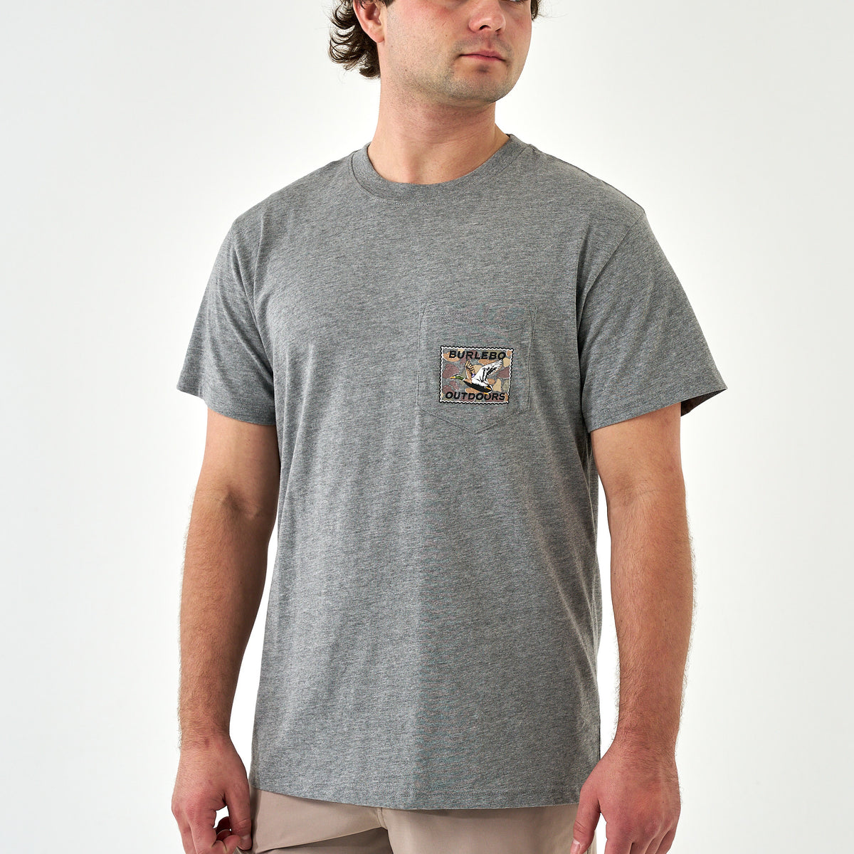 Model wearing dark heather gray Burlebo Outdoors pocket t-shirt with duck stamp graphic, front view.