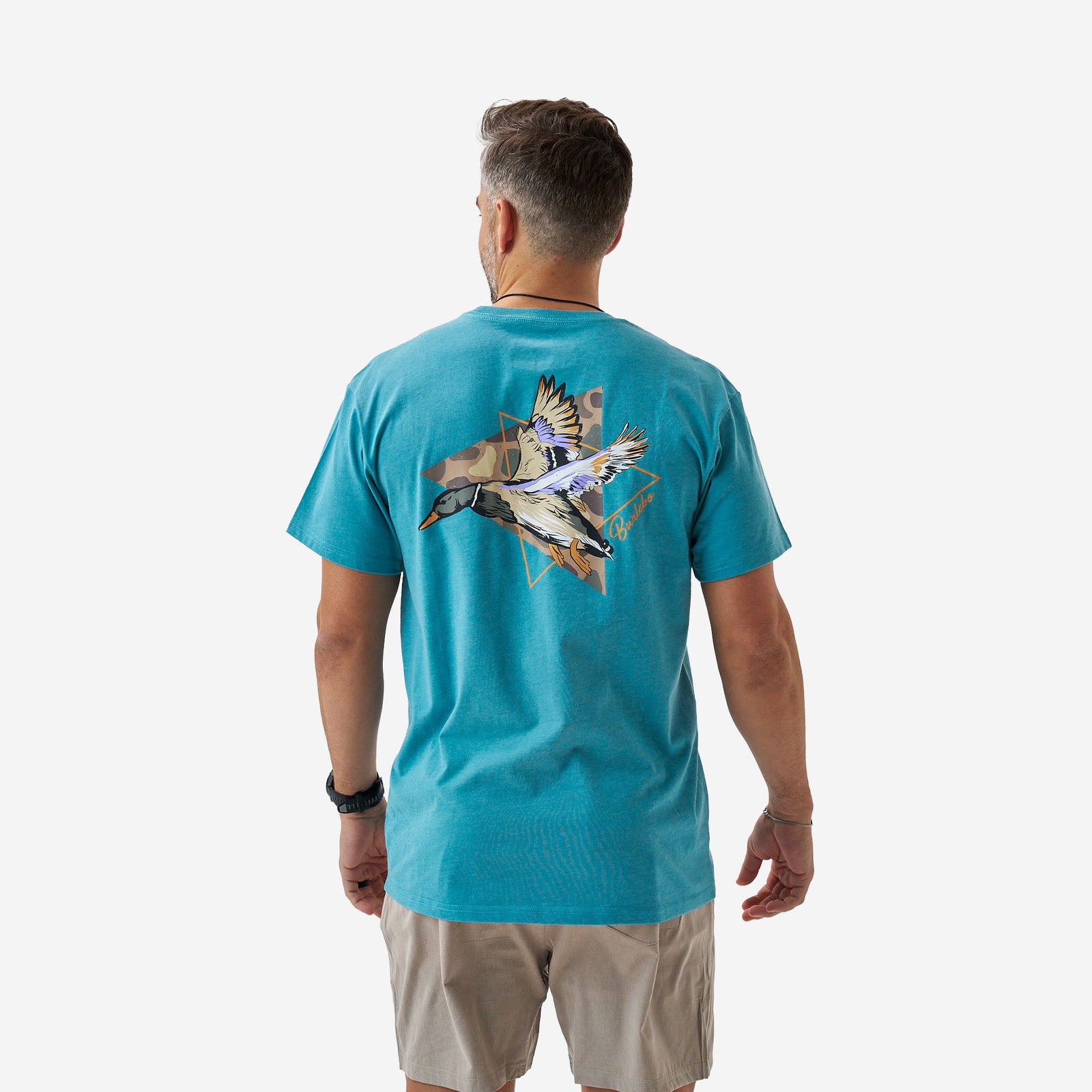Rear full-body view of a male model wearing a teal short sleeve Burlebo t-shirt with a large flying mallard duck graphic over geometric shapes on the back.