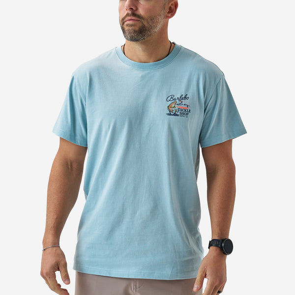 Front view of a male model wearing a light blue Burlebo short sleeve t-shirt with a small “Burlebo Tackle Shop Supply Co.” chest graphic featuring a trout and fly fishing design.