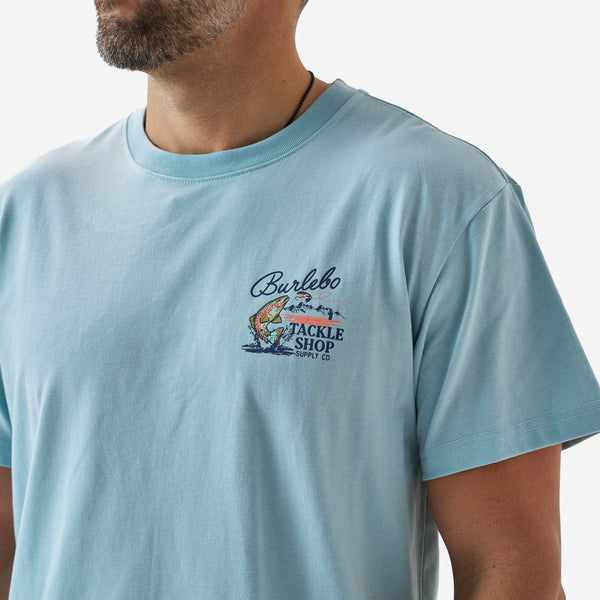 Close-up of the chest graphic on a light blue Burlebo t-shirt showing a trout, fly lure, and “Burlebo Tackle Shop Supply Co.” text.