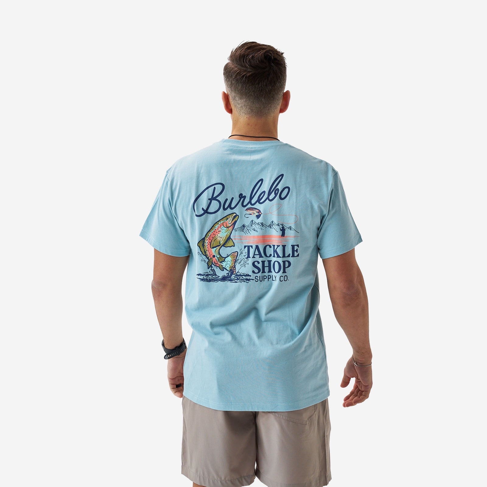 Rear full-body view of a male model wearing a light blue Burlebo short sleeve t-shirt featuring a large “Burlebo Tackle Shop Supply Co.” graphic with a jumping trout, fly lure, and mountain scene.