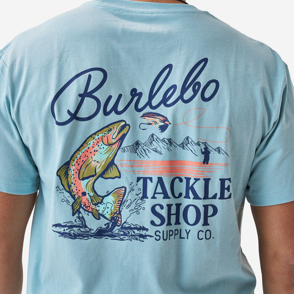 Close-up of the back graphic on a light blue Burlebo t-shirt showing a colorful trout jumping from water with “Burlebo Tackle Shop Supply Co.” text and fly fishing illustration.