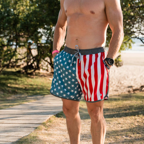 Burlebo Men's Swim Trunks in Throwback USA. Sizes S-XXL. Man standing outdoors wearing trunks with patriotic stars and stripes design, hands in pockets, with beach background.