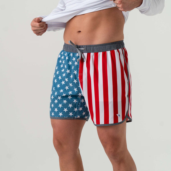 Burlebo Men's Swim Trunks in Throwback USA. Sizes S-XXL. Side view of a man lifting his shirt to show the star-and-stripe design trunks with a grey waistband and drawstring.