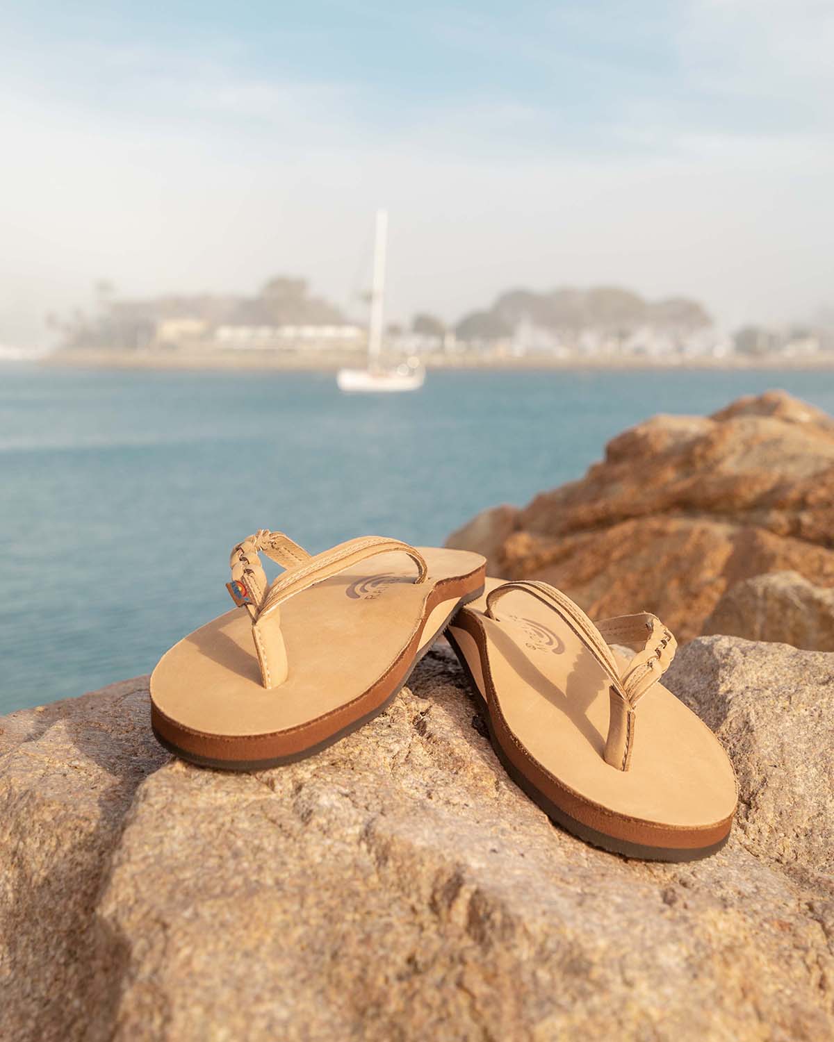 Tan leather flip-flop sandals resting on coastal rocks with ocean water and a sailboat in the background.
