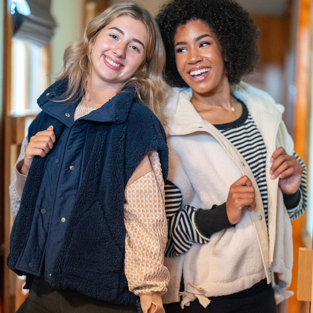 Two women standing together, smiling, with one wearing a navy blue vest and the other in a cream vest.