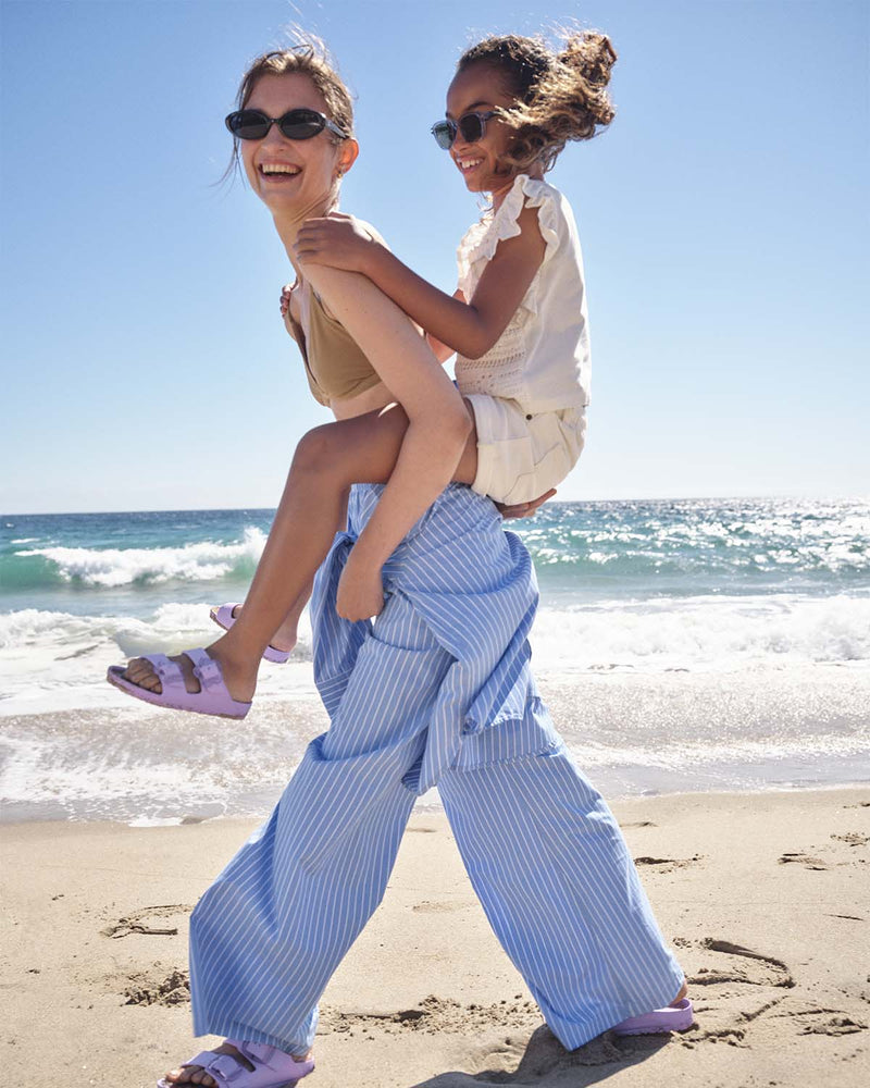 A mom and daughter walking across the beach in summer clothing. They are wearing matching light purple Birkenstock sandals.
