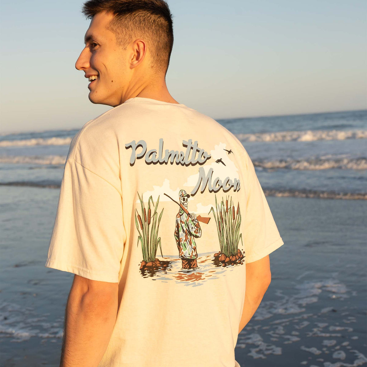Man wearing a 'Palmetto Moon' t-shirt with a fishing design on a beach.