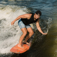 Man wake surfing on a lake wearing a black life vest and patterned swim trunks on an orange surfboard.