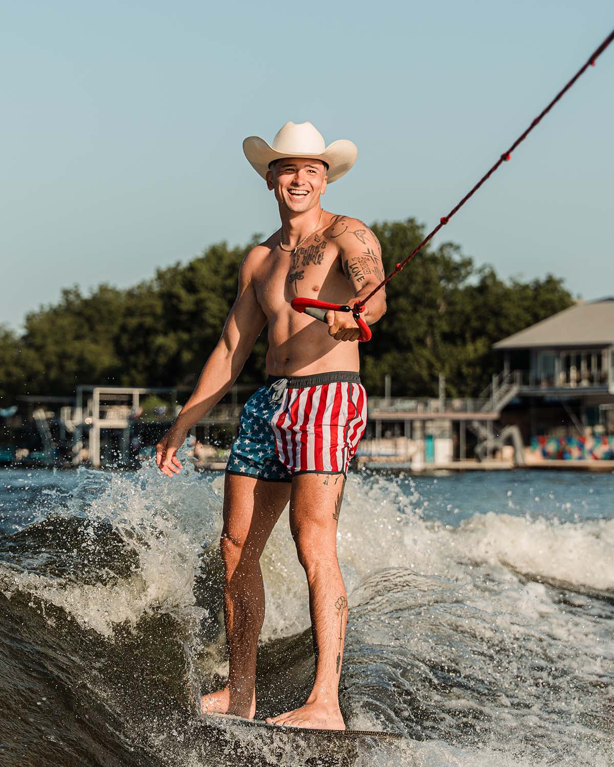 A man wakeboarding in a lake. He is wearing American flag printed swim trunks and a cowboy hat.