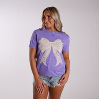 Women’s lavender short sleeve graphic t-shirt with large lace bow design on front, styled with denim shorts in studio setting.