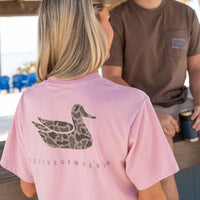 Woman wearing a pink t-shirt with camo duck graphic on back standing near a beachside bar.