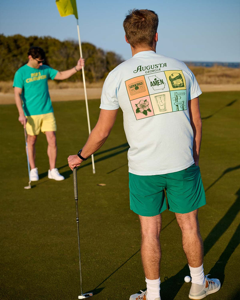 Man wearing light blue Augusta Georgia graphic T-shirt and green shorts holding a putter on a golf course green with flag in background.