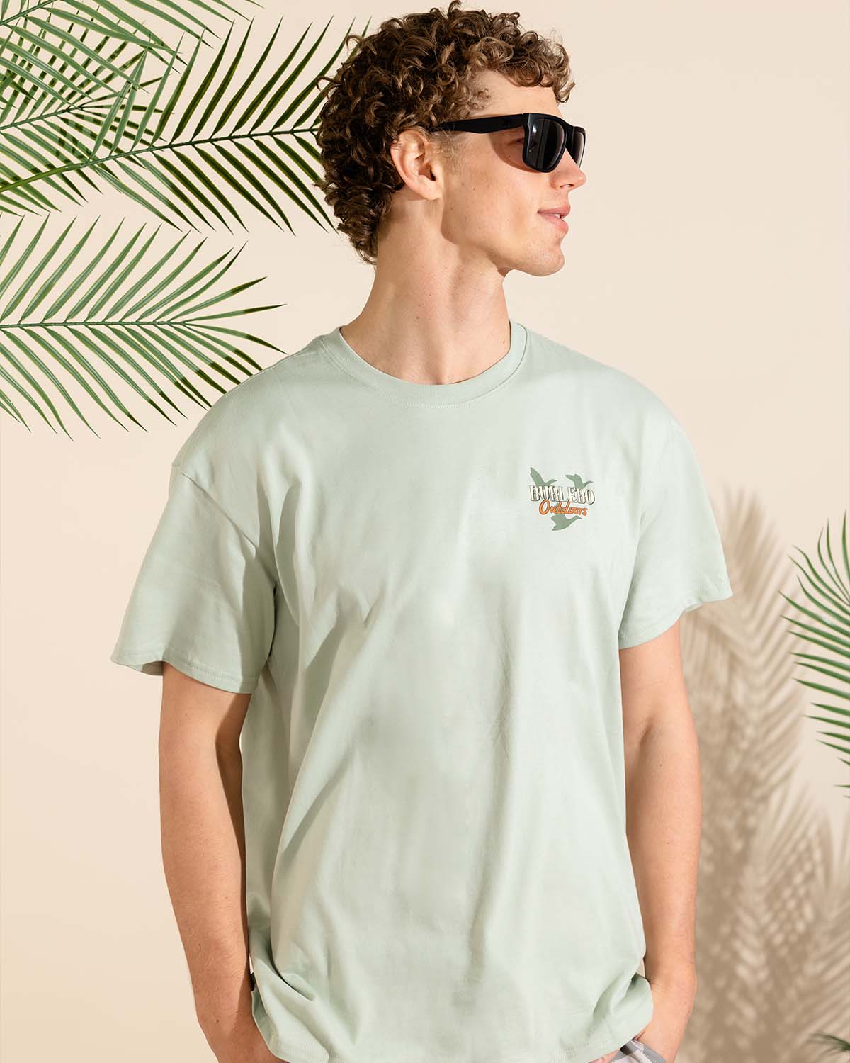 Male model wearing sage green graphic T-shirt and black sunglasses standing in front of palm leaves against a neutral background.