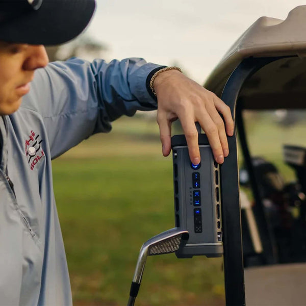 A man places a gray Turtlebox Ranger speaker on the metal bar of a golf cart, showing its magnetic mount and illuminated control panel.
