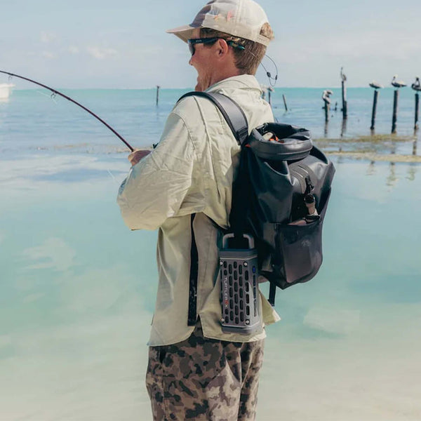 A man stands on the beach fishing with a black backpack that holds a gray Turtlebox Ranger speaker clipped to the side.