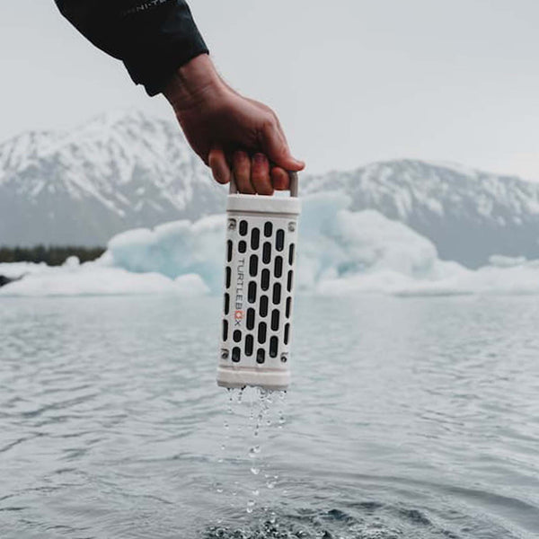 A person holds a white Turtlebox Ranger speaker dripping with water above a cold lake, with snowy mountains and glaciers in the background.