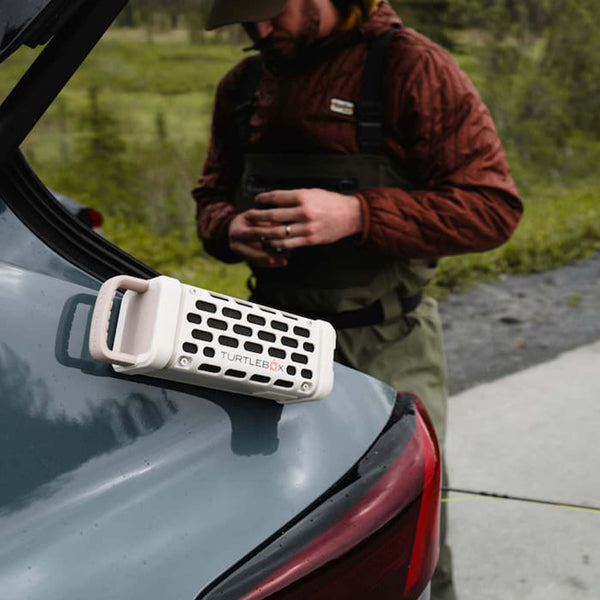 Ivory Turtlebox Ranger speaker magnetically mounted to the back of a car, while a man in outdoor gear stands nearby adjusting fishing gear.