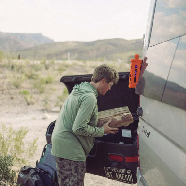 A man unloads firewood from a car near a van in a desert setting with an orange Turtlebox Ranger speaker magnetically mounted to the van.
