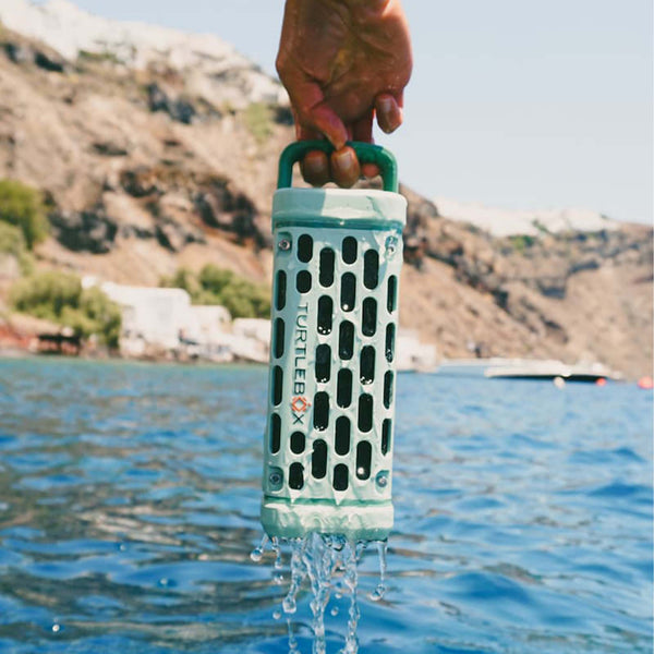 A hand holds a dripping River Rock Turtlebox Ranger speaker above blue water with cliffs and a boat in the background.