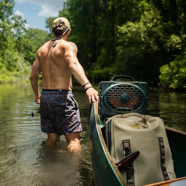 Turtlebox Gen 3 Green Portable Speaker sitting inside a canoe as a man stands beside it in a shallow creek, surrounded by trees.