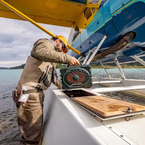 Turtlebox Gen 3 Green Portable Speaker being lifted by a man into a storage compartment on a docked floatplane at the water’s edge.