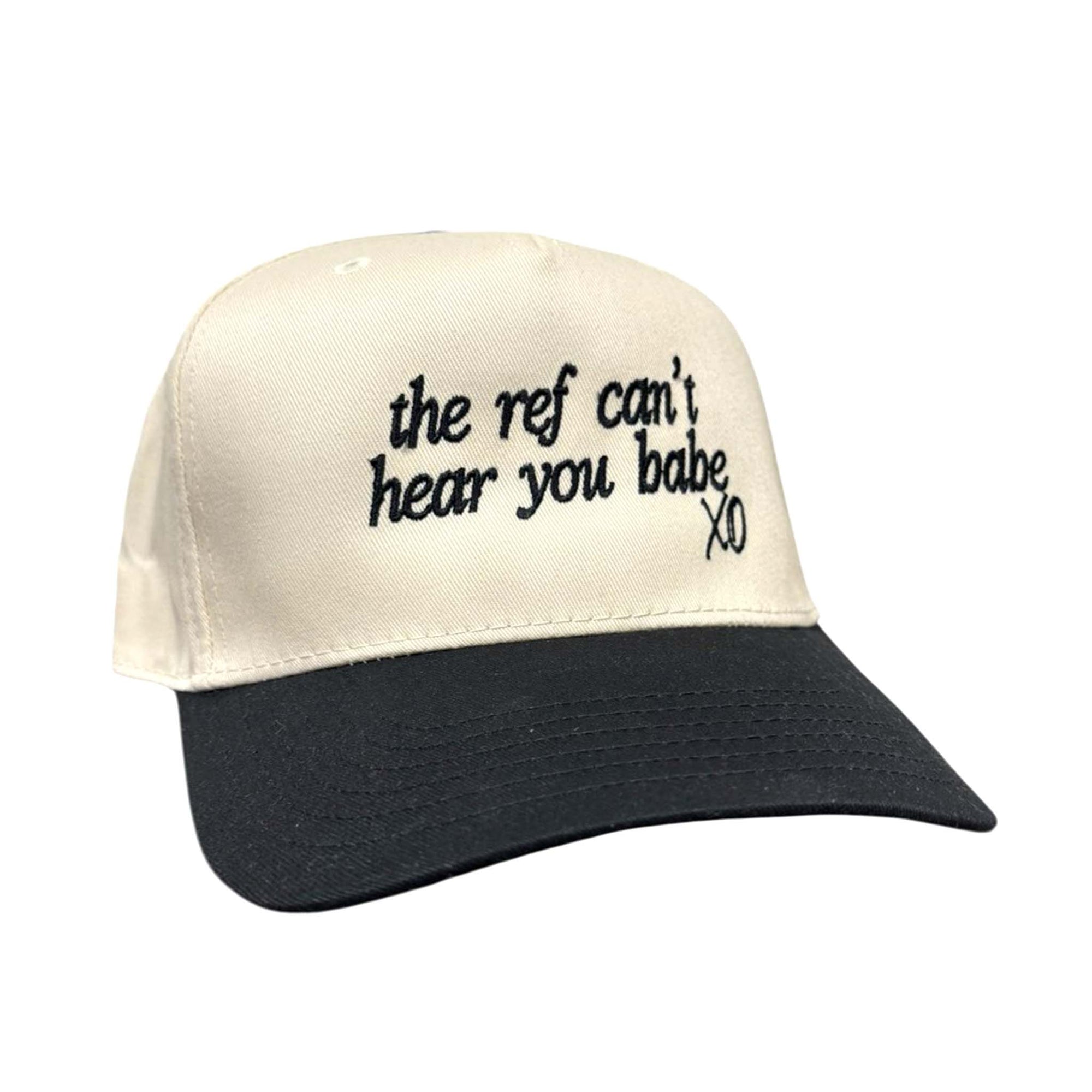 Cream and black baseball cap with embroidered phrase “the ref can’t hear you babe XO” on the front and adjustable back closure.
