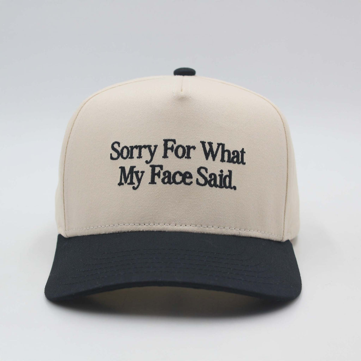 Beige and black baseball hat by The Happiest with the embroidered phrase ‘Sorry For What My Face Said.’ displayed on the front panel.