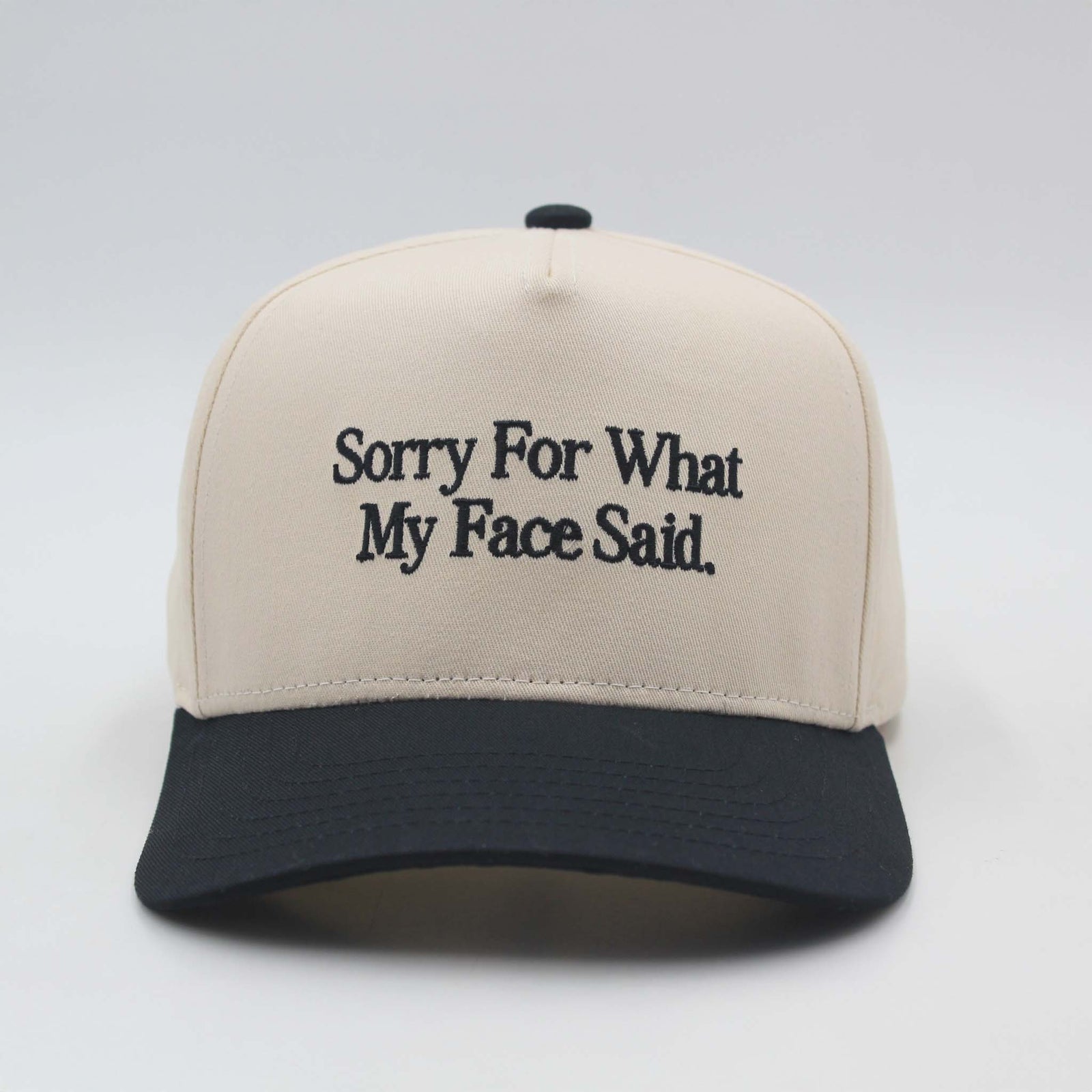 Beige and black baseball hat by The Happiest with the embroidered phrase ‘Sorry For What My Face Said.’ displayed on the front panel.