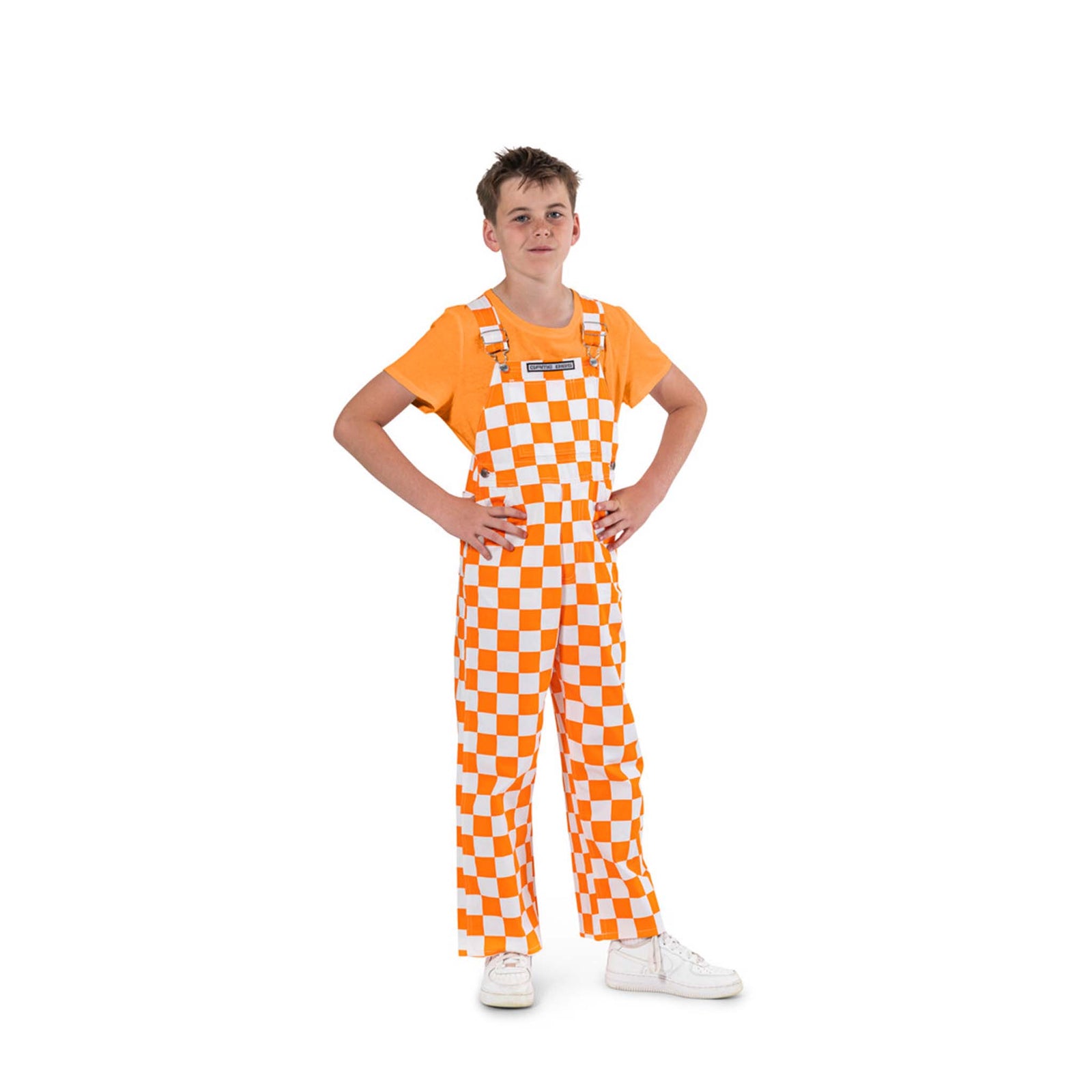 Young boy wearing orange and white checkered overall bibs paired with an orange short sleeve shirt and white tennis shoes.