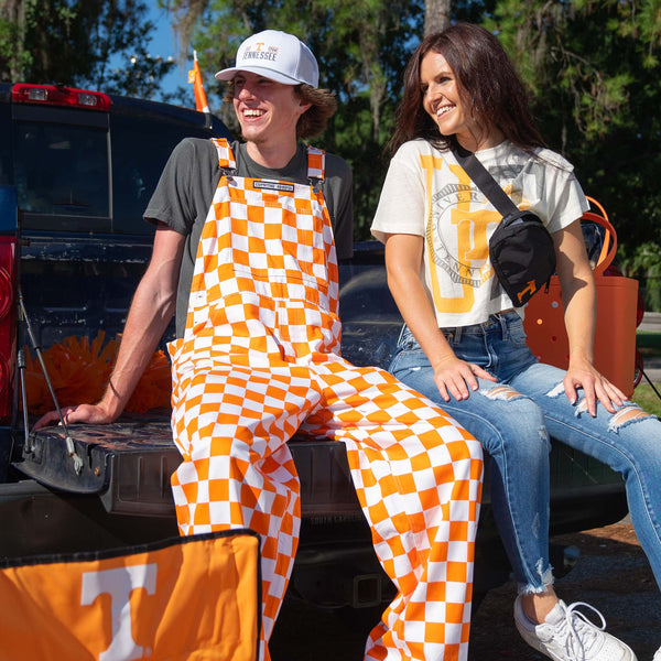 Man wearing orange and white checkered overalls and Tennessee hat sits on a truck tailgate next to woman in Tennessee tee, both smiling at a tailgate.