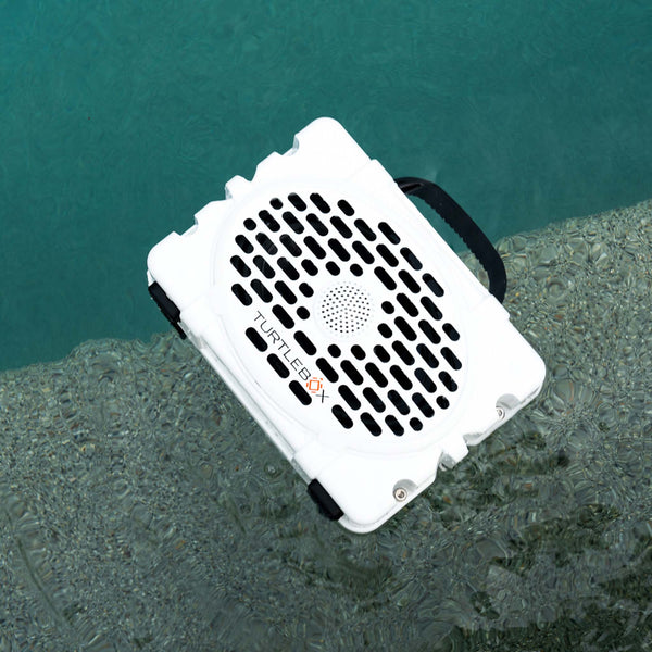 A white Turtlebox portable speaker rests at the edge of a pool, partially in the water. The speaker has a black grille, carrying handle, and corner bumpers, showcasing its rugged, waterproof design against the clear blue pool water.
