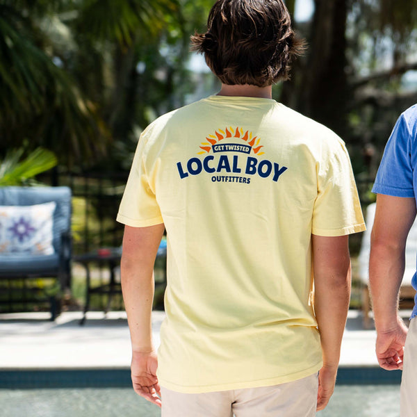 A man stands with his back to the camera wearing a pale yellow Local Boy Outfitters t-shirt. The back of the shirt features a sun graphic and the phrase “Get Twisted” above the bold navy text “Local Boy Outfitters.” He is standing next to another person in a blue shirt, near a poolside area with outdoor furniture and palm trees in the background.