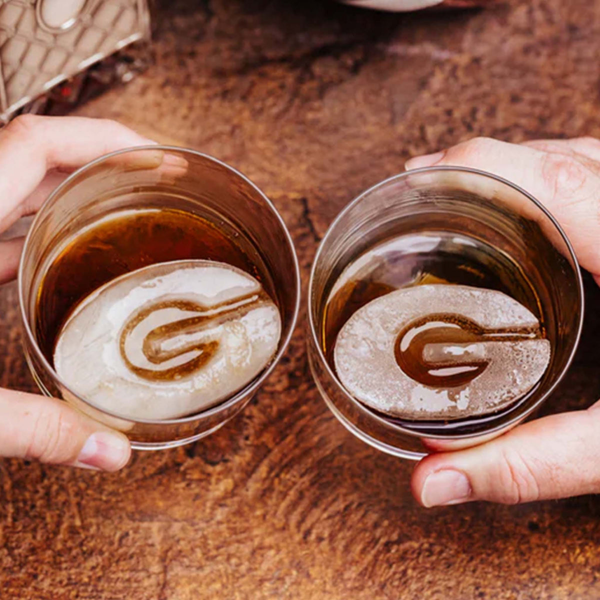 Two drink glasses with G-shaped ice cubes held in hands on wooden table