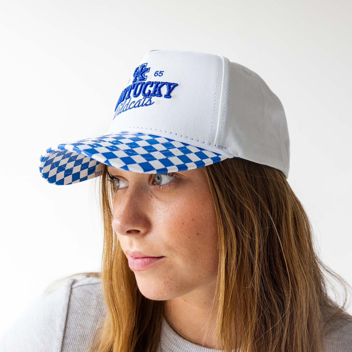 Kentucky Wildcats white cap with embroidered logo and “Wildcats” script, featuring a blue and white checkered brim, worn by a model facing left.
