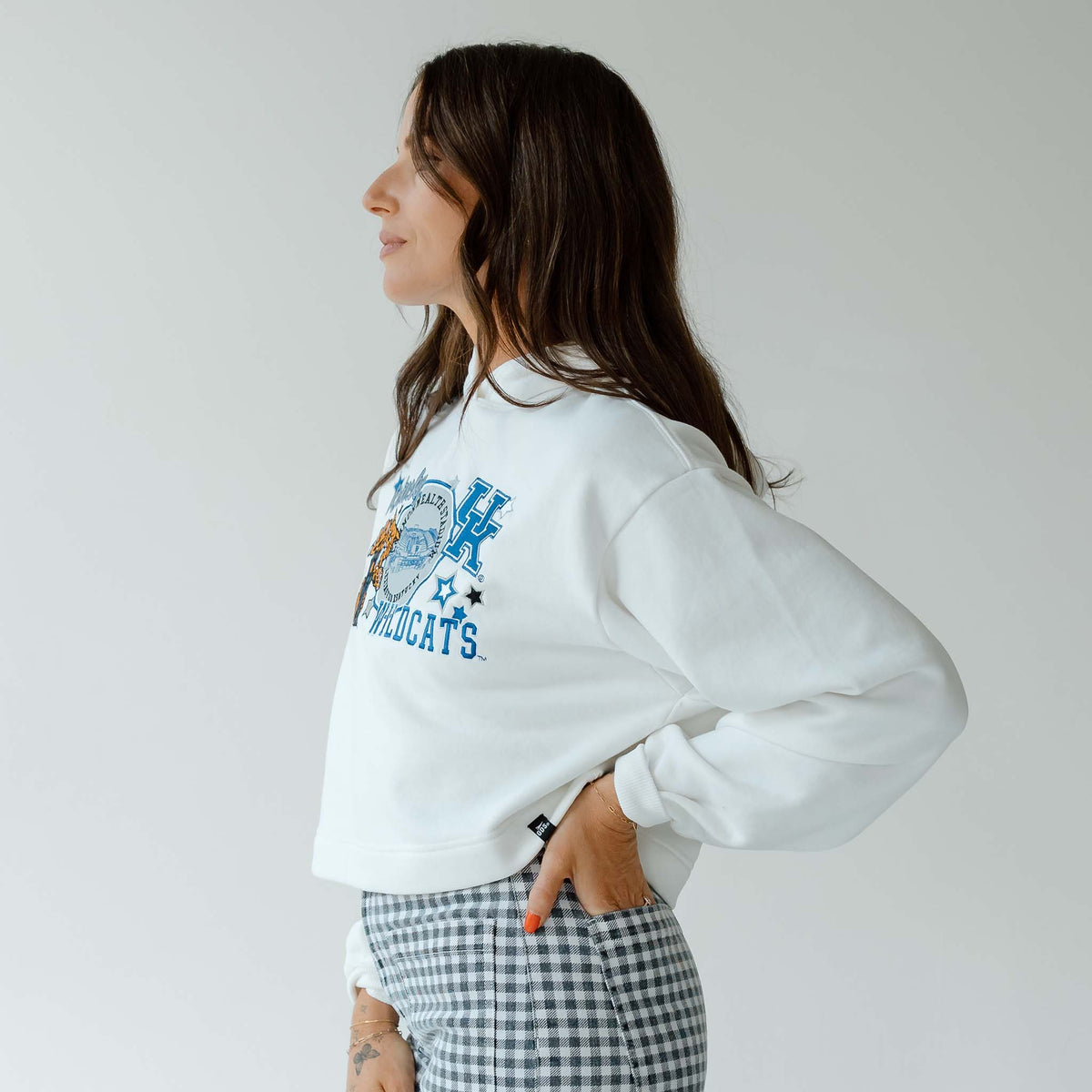 Side view of woman wearing a white cropped hoodie with “Kentucky Wildcats” graphic and gingham pants.