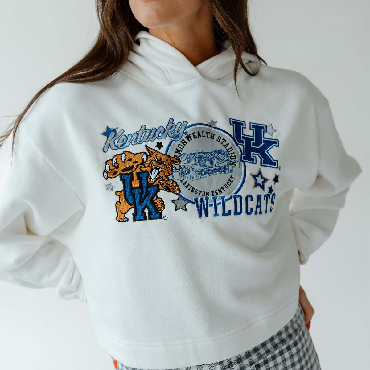 Close-up of white cropped hoodie with “Kentucky Wildcats” graphic featuring mascot, stadium, and stars, paired with gingham pants.