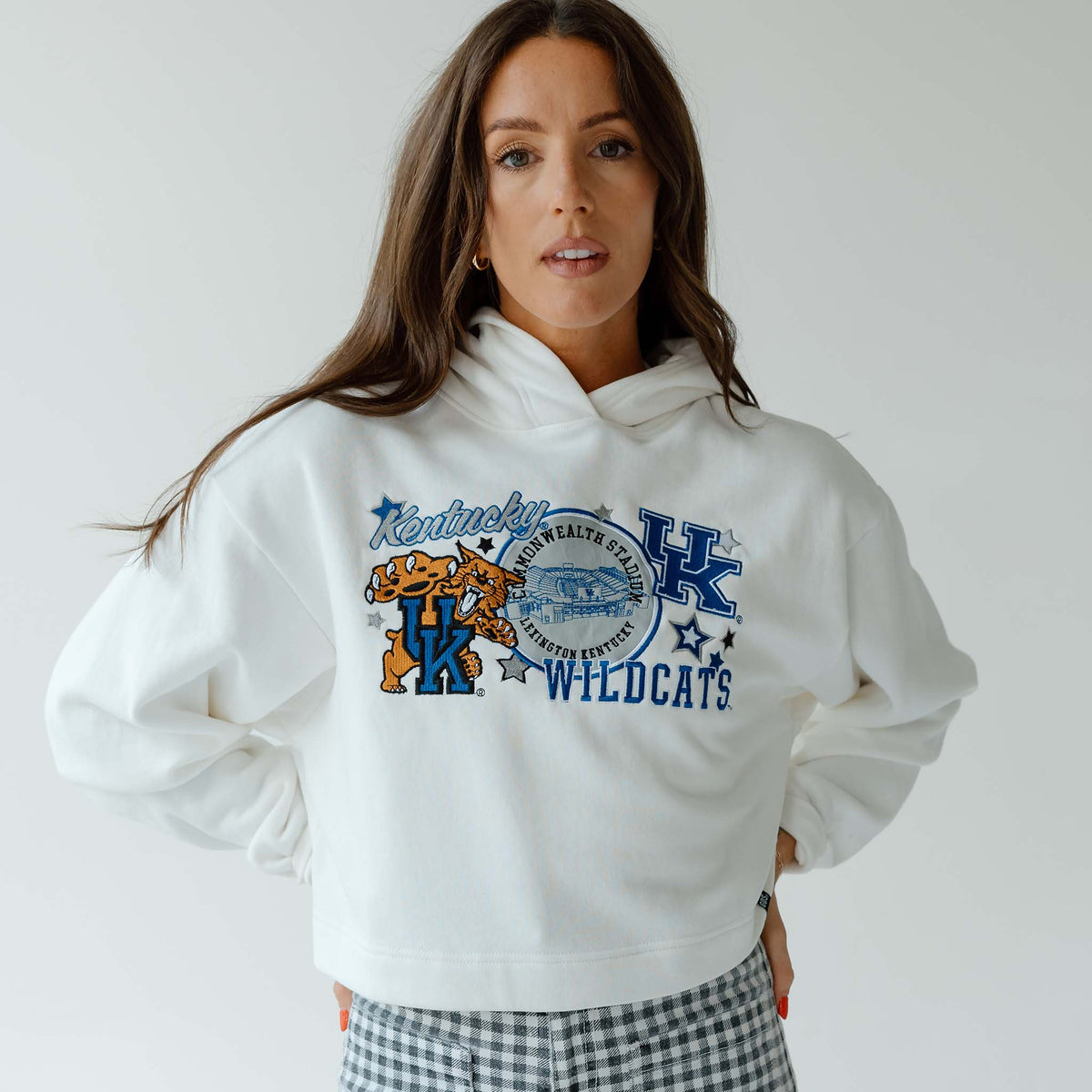 Woman facing forward wearing a white cropped hoodie with “Kentucky Wildcats” graphic and gingham pants against a light gray background.