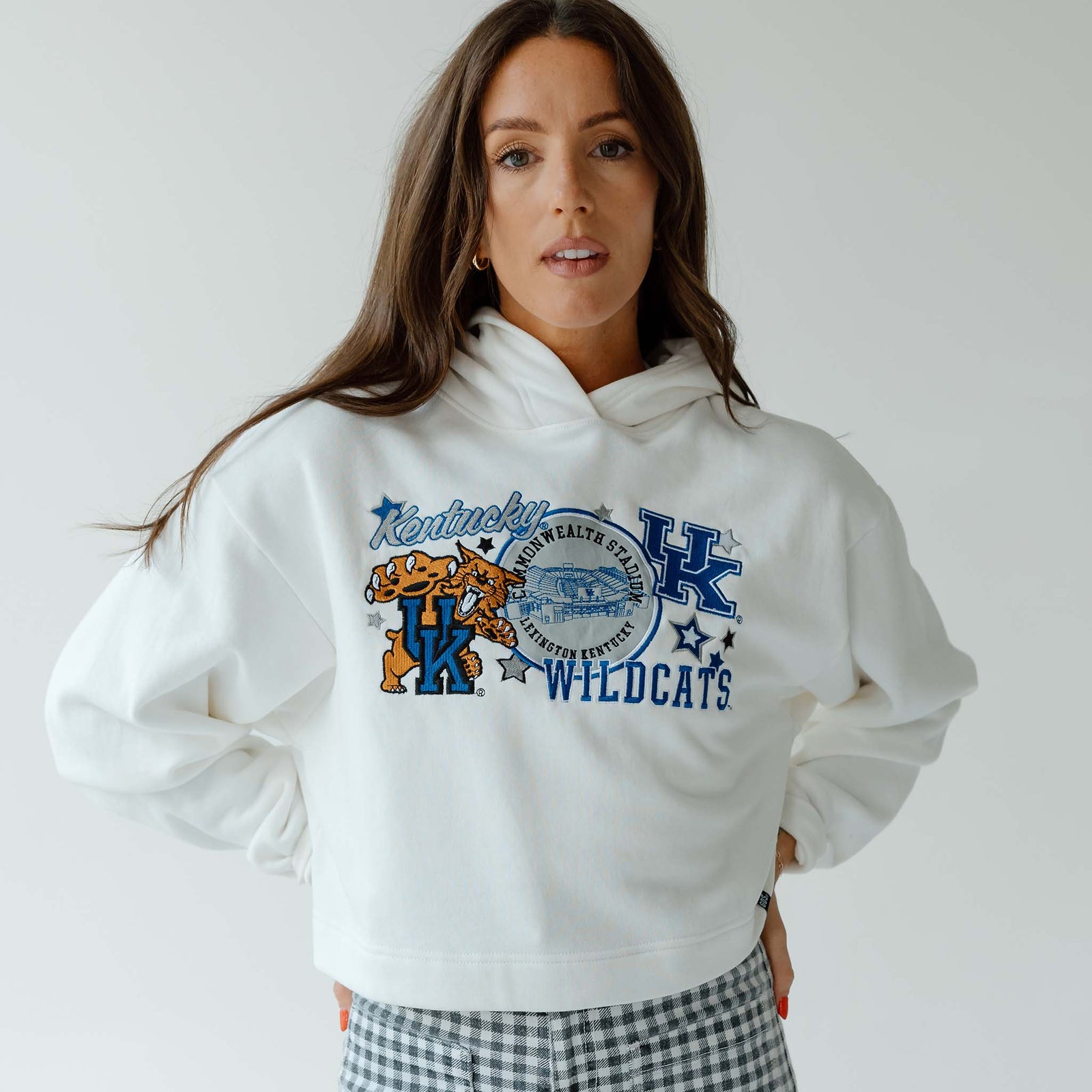 Woman facing forward wearing a white cropped hoodie with “Kentucky Wildcats” graphic and gingham pants against a light gray background.