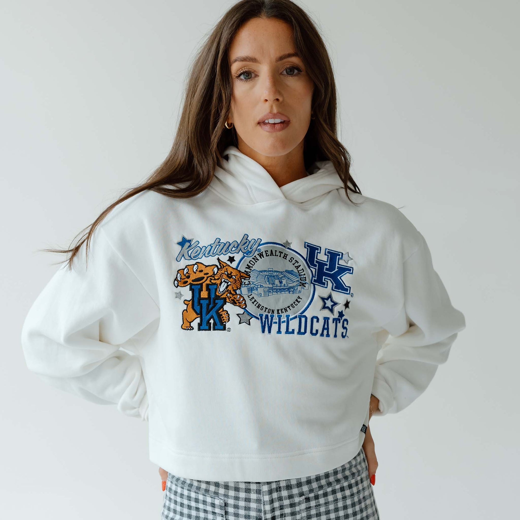 Woman facing forward wearing a white cropped hoodie with “Kentucky Wildcats” graphic and gingham pants against a light gray background.