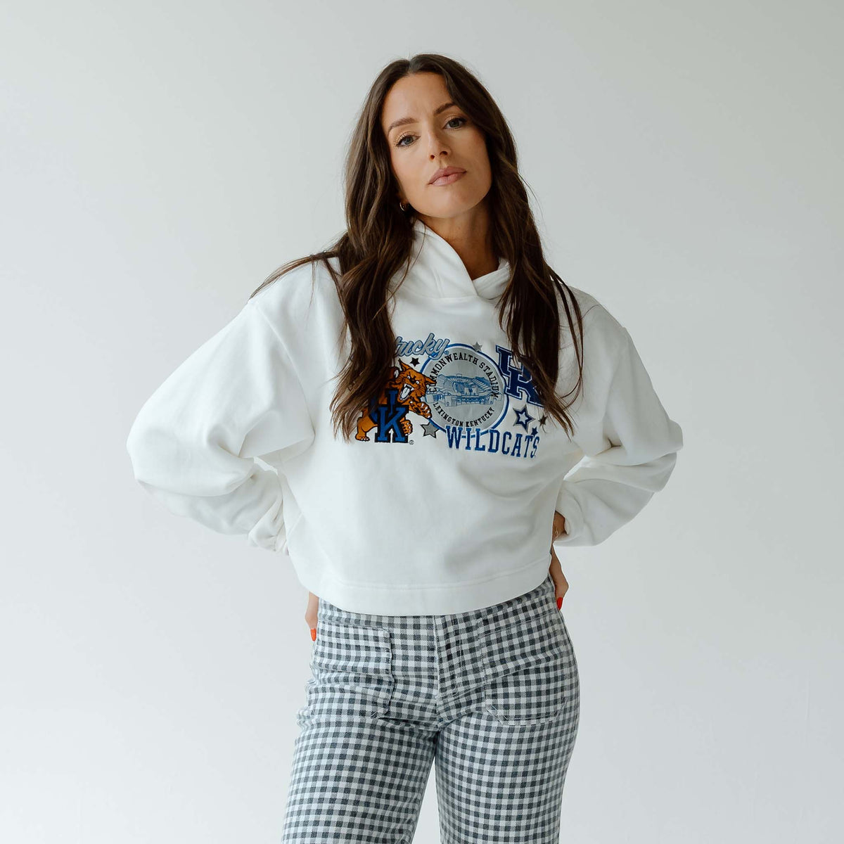 Woman wearing a white cropped hoodie with “Kentucky Wildcats” graphic and gingham pants, standing with hands on hips against a light gray background.