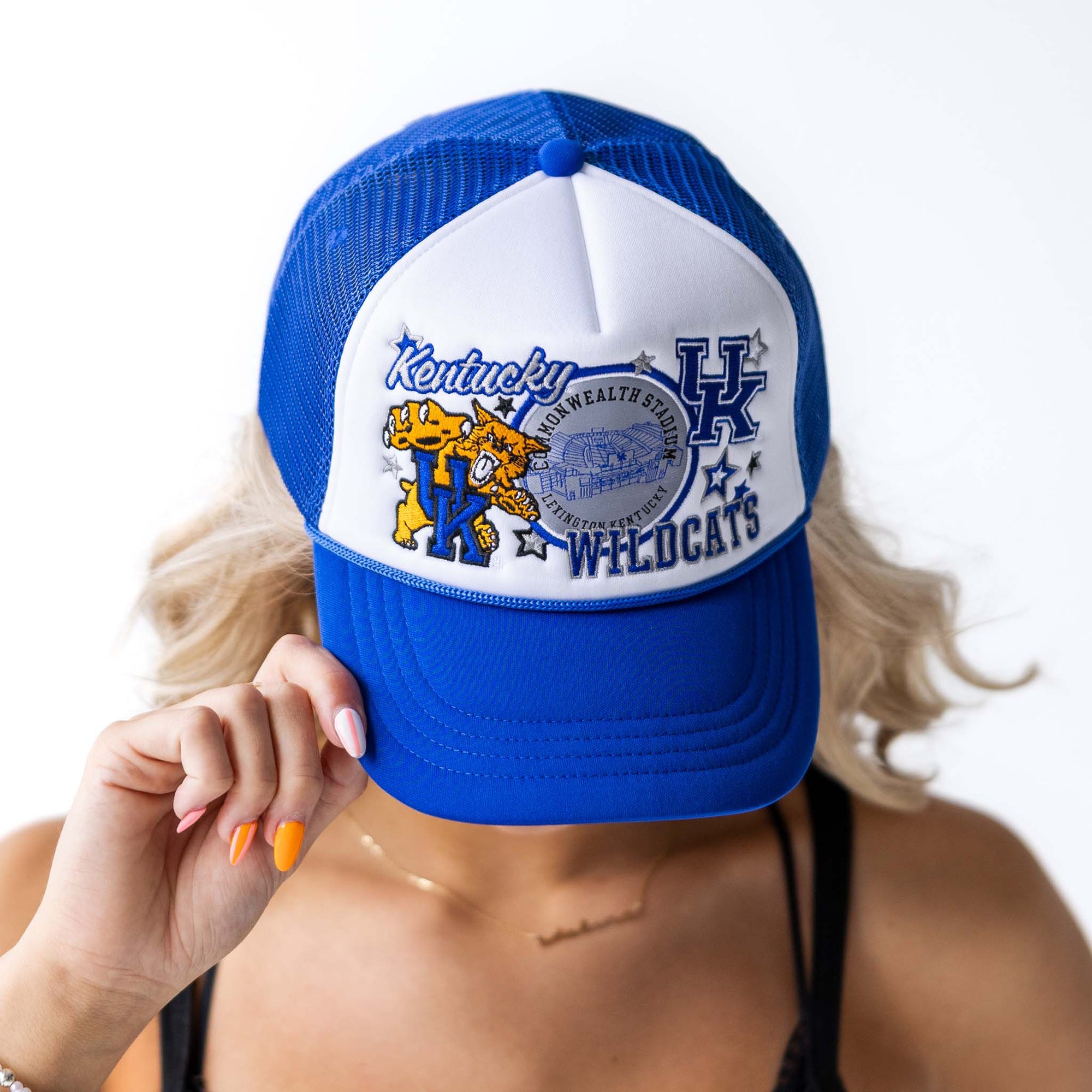 Woman wearing a royal blue and white Kentucky Wildcats trucker hat with mascot, stadium, and logo graphic, holding brim with one hand.