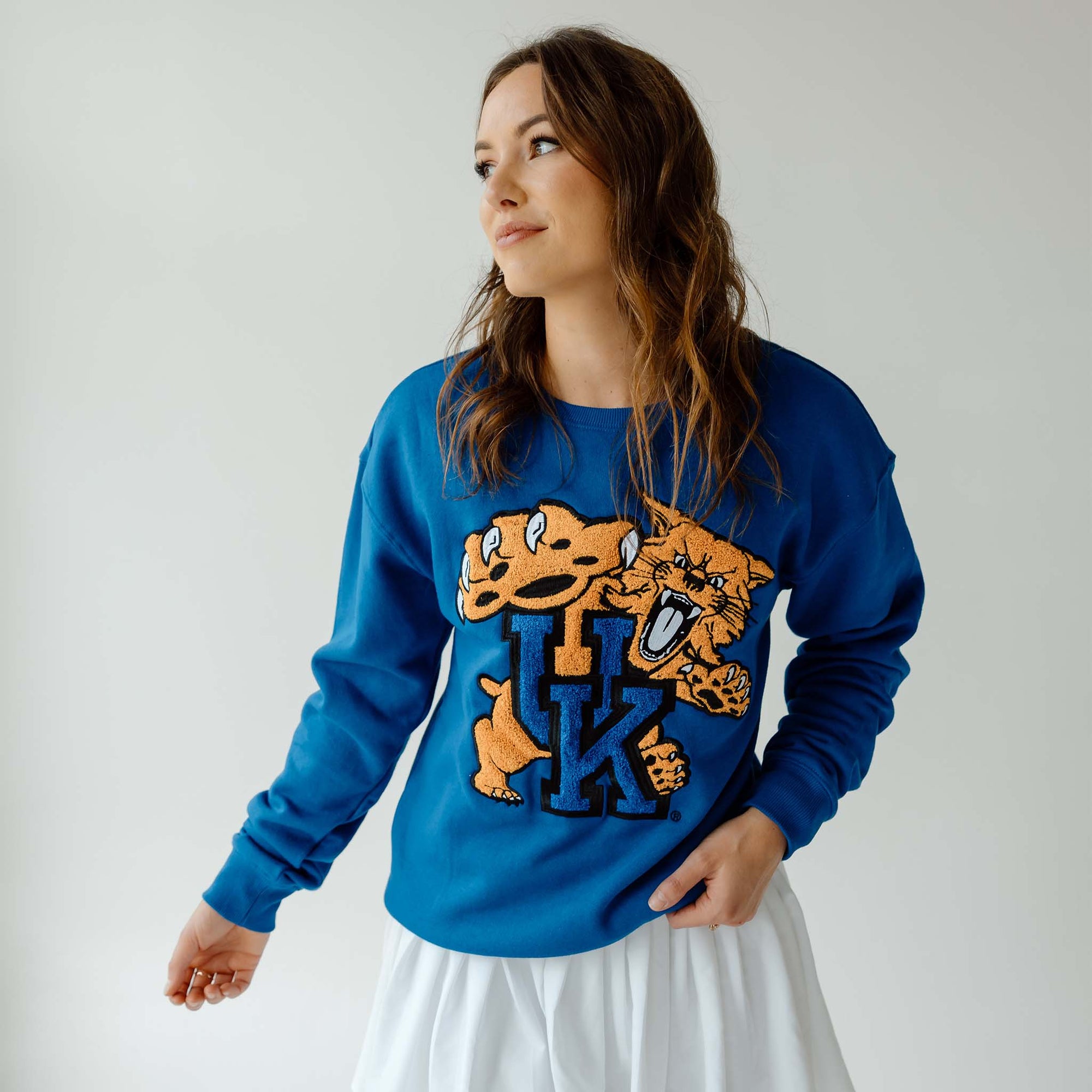 Woman wearing royal blue crewneck sweatshirt with large Kentucky Wildcats chenille patch logo, looking to the side against light gray background.