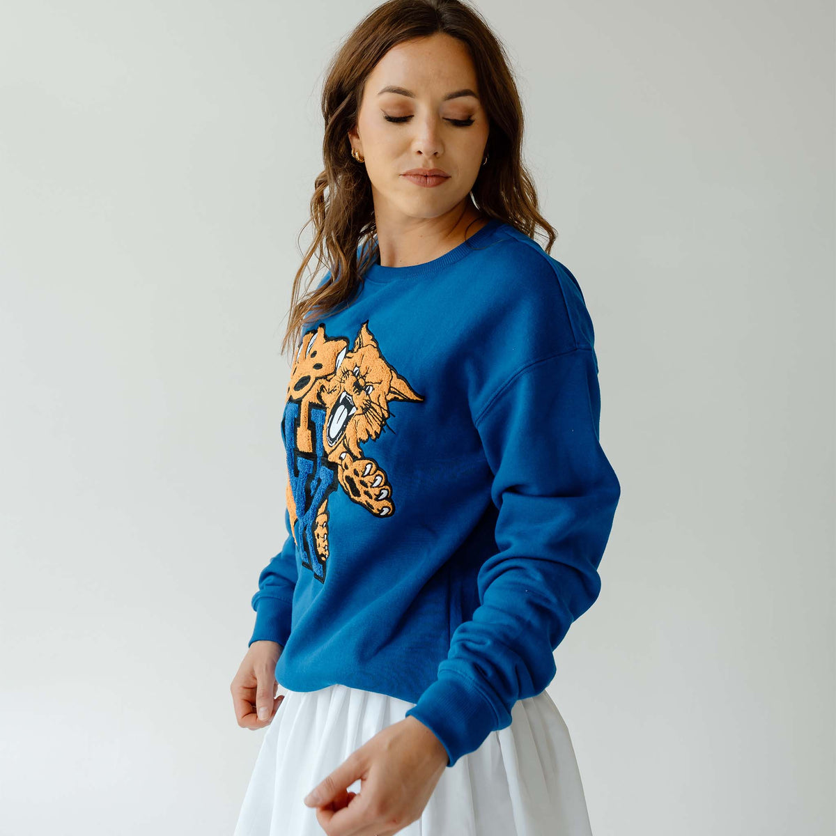 Woman wearing royal blue crewneck sweatshirt with large Kentucky Wildcats chenille patch logo, standing against light gray background.