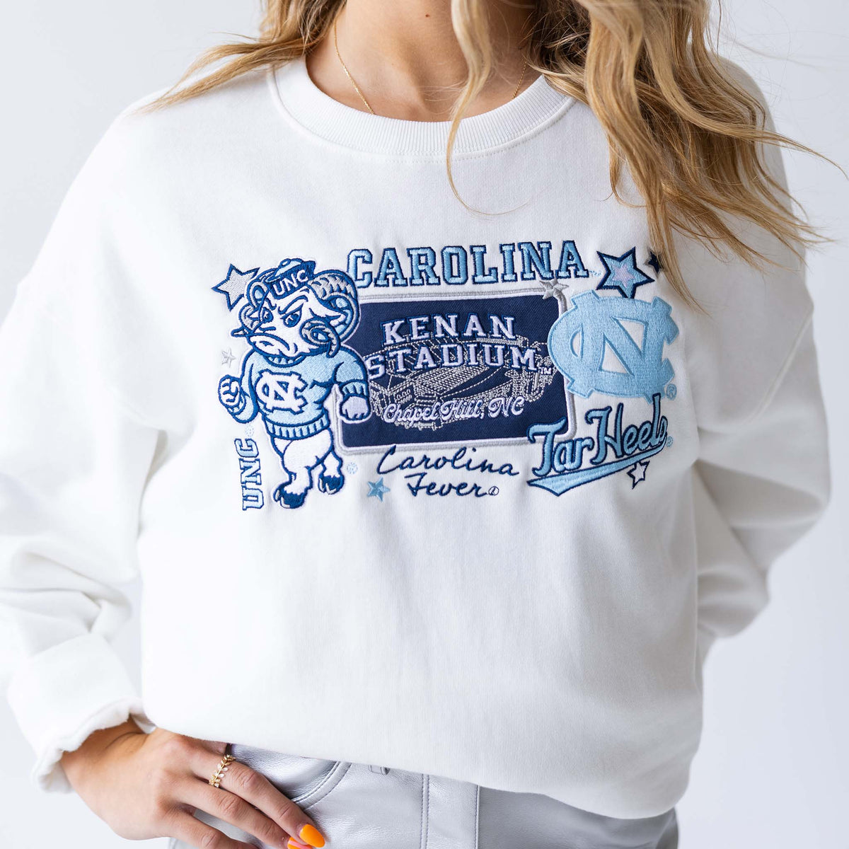 Close-up of woman wearing white crewneck sweatshirt with embroidered UNC mascot, Kenan Stadium, and team logos.
