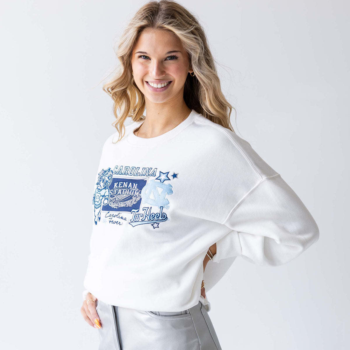 Woman smiling in white crewneck sweatshirt with embroidered UNC mascot, Kenan Stadium, and team logos, hands on hips.