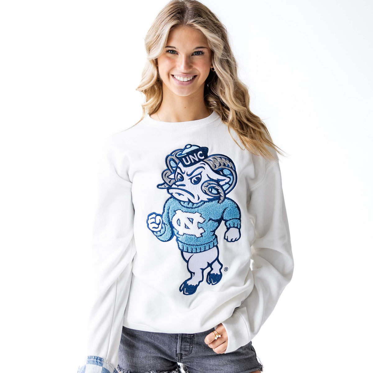 Woman smiling in white crewneck sweatshirt with oversized chenille UNC Rameses mascot patch, paired with frayed gray shorts.