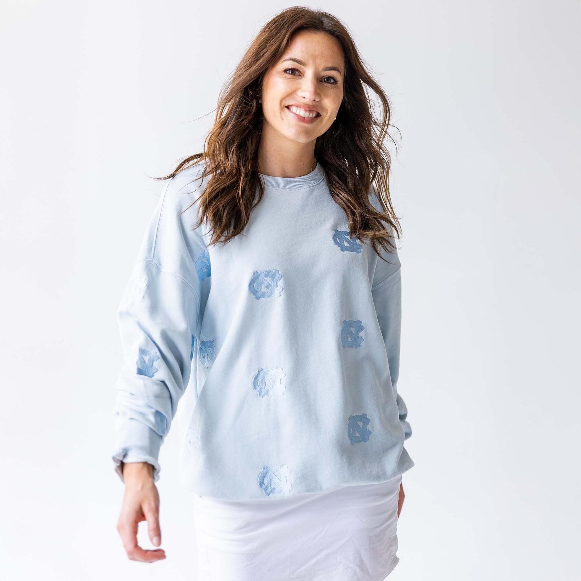 Woman smiling in light blue crewneck sweatshirt with allover tonal embroidered UNC logos, paired with white skirt.