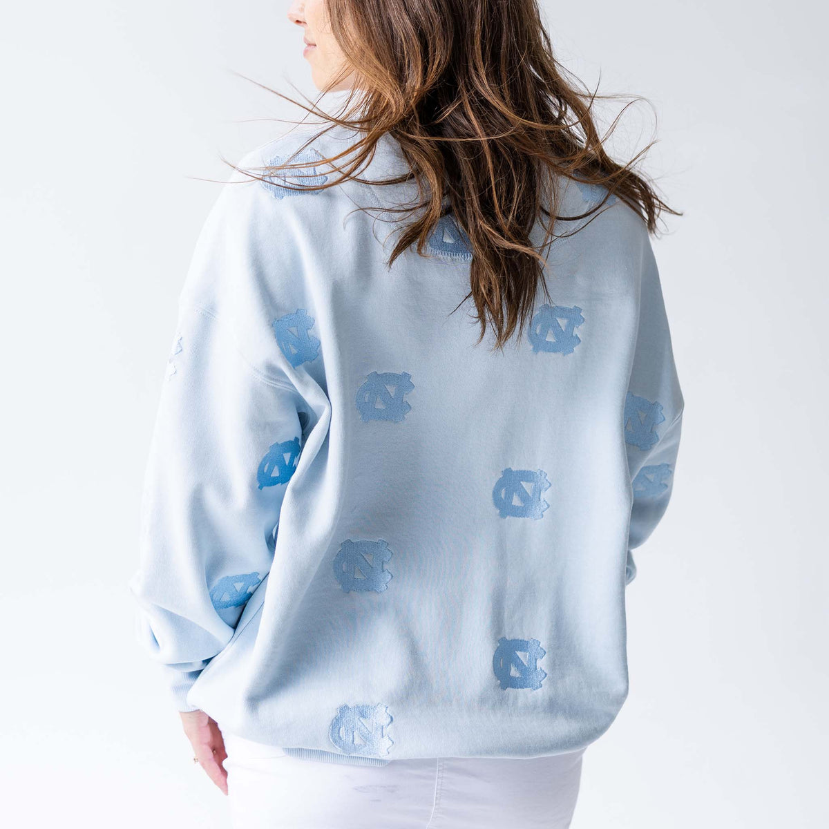 Back view of woman wearing light blue crewneck sweatshirt with allover tonal embroidered UNC logos, paired with white skirt.