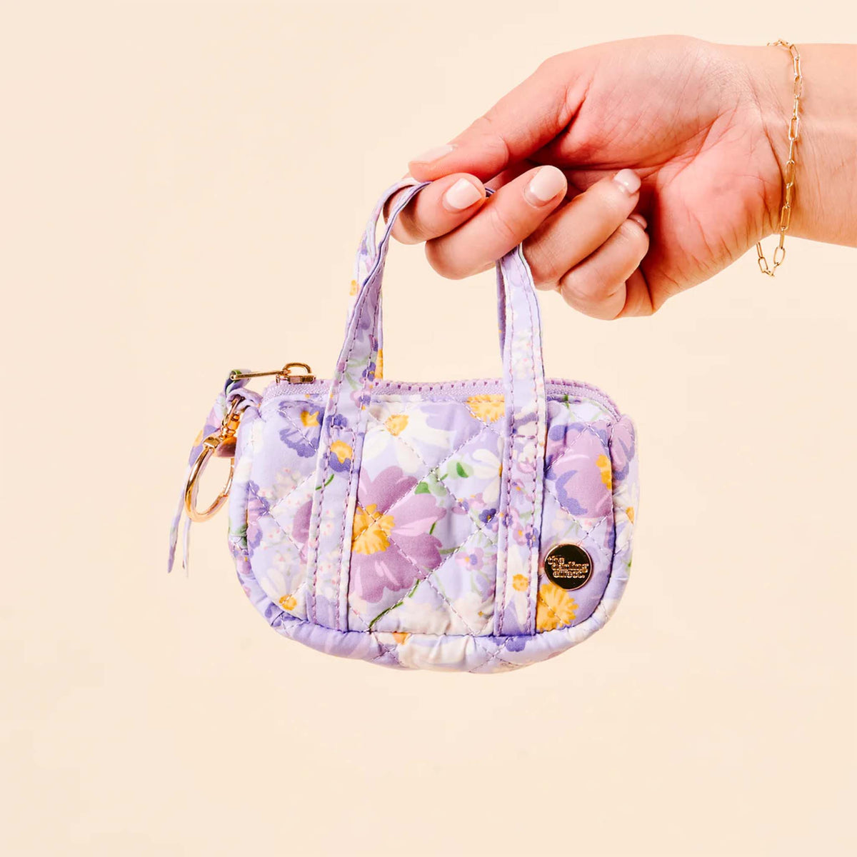 Hand holding the purple floral mini duffle keychain, showing front design with gold logo charm and top handles.